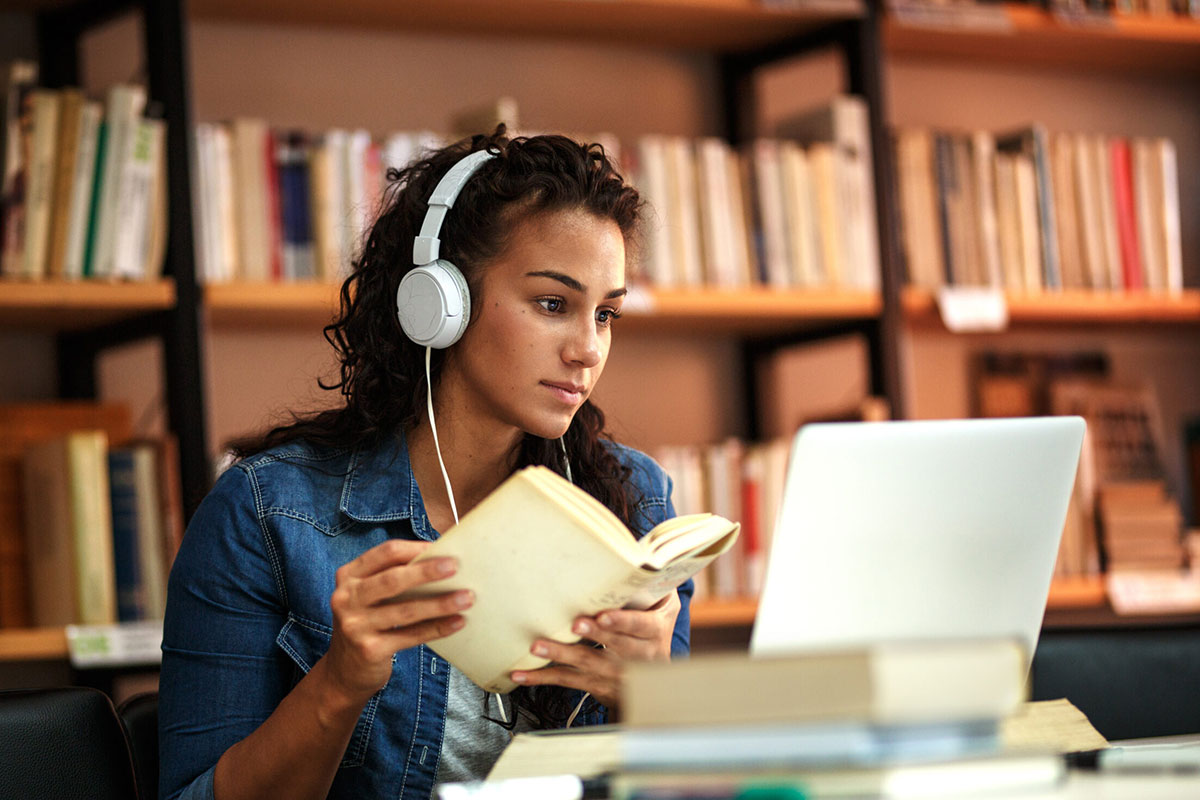 Female student in library, listening to music, studying on her computer, book in hand