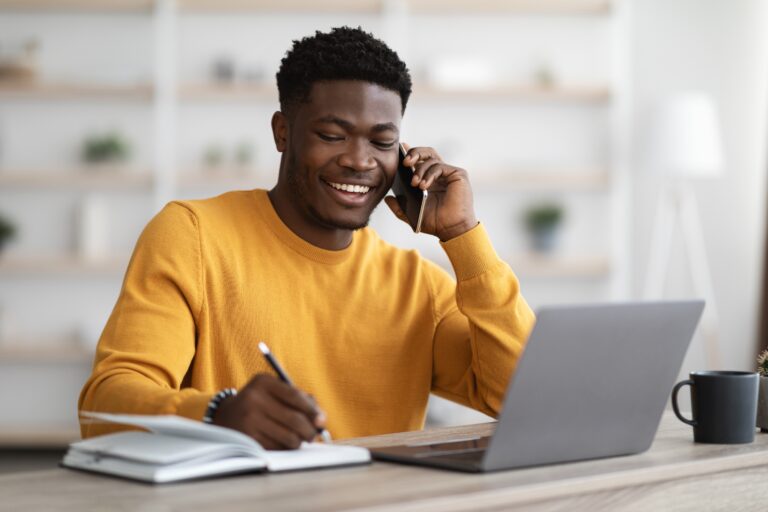 Happy african american young man freelancer or businessman working from home, sitting at table in front of modern laptop, having phone conversation, taking notes and smiling